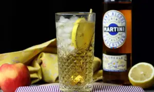 Orzeźwiający spritz bezalkoholowy z plasterkiem cytryny w szklance z lodem. Obok butelka Martini Floreale i jabłko.