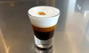 Kawa espresso corretto z pianką i kroplą likieru, podana w szklance na metalowym blacie.