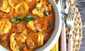 Pyszne tortellini z mięsem w kremowym sosie pomidorowym z warzywami, udekorowane listkiem bazylii. Obok widelec i łyżka.