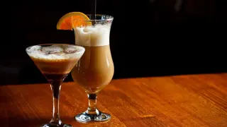 Dwa drinki na drewnianym stole: w kieliszku martini espresso martini, a w wysokiej szklance kawa z alkoholem jak się nazywa, ozdobiona plasterkiem pomarańczy.