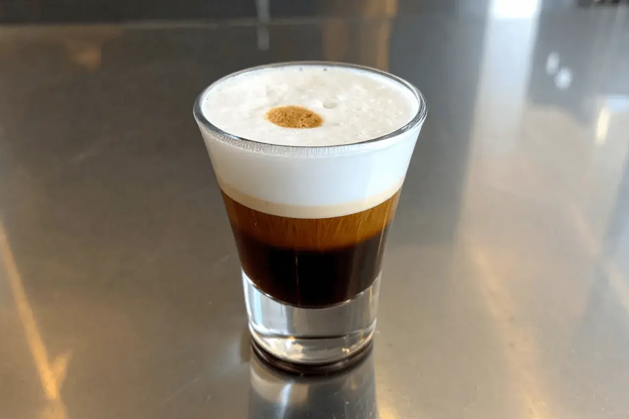 Kawa espresso corretto z pianką i kroplą likieru, podana w szklance na metalowym blacie.