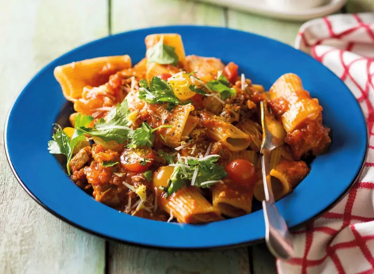 Pyszne mezzi rigatoni w sosie pomidorowym z mięsem mielonym, pomidorkami koktajlowymi i świeżą natką pietruszki, podane na niebieskim talerzu.