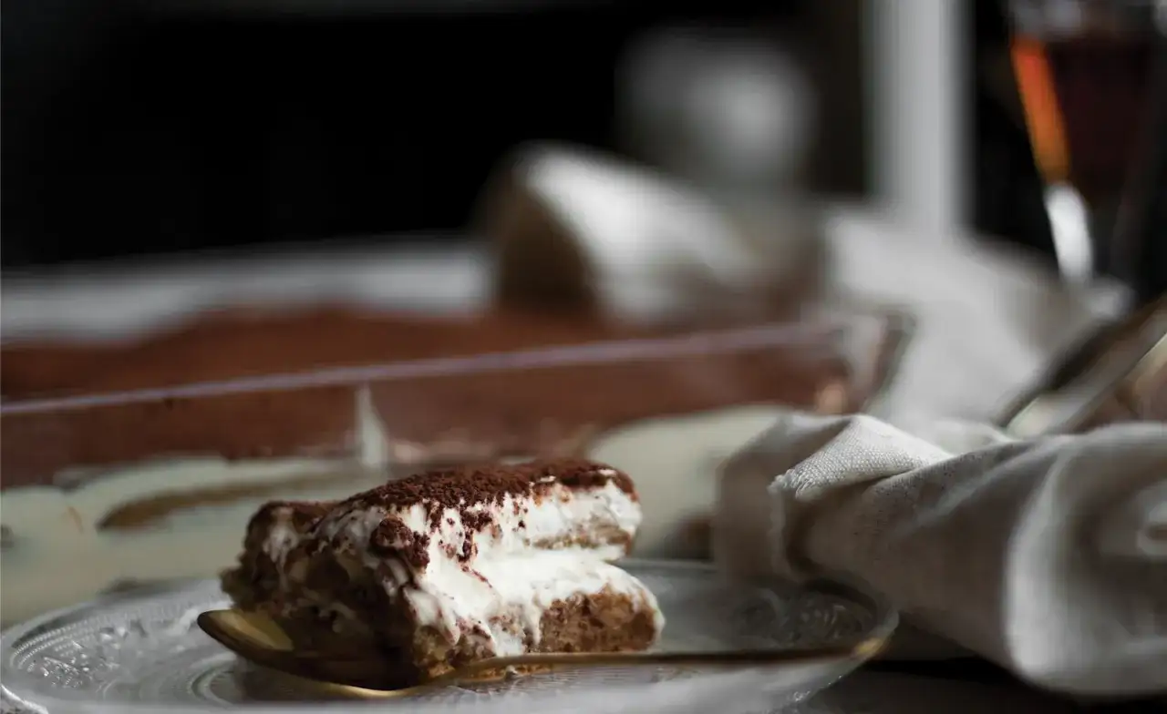 Pyszne tiramisu na talerzyku, z widocznym w tle deserem w naczyniu. Zastanawiasz się, jaki alkohol do tiramisu?