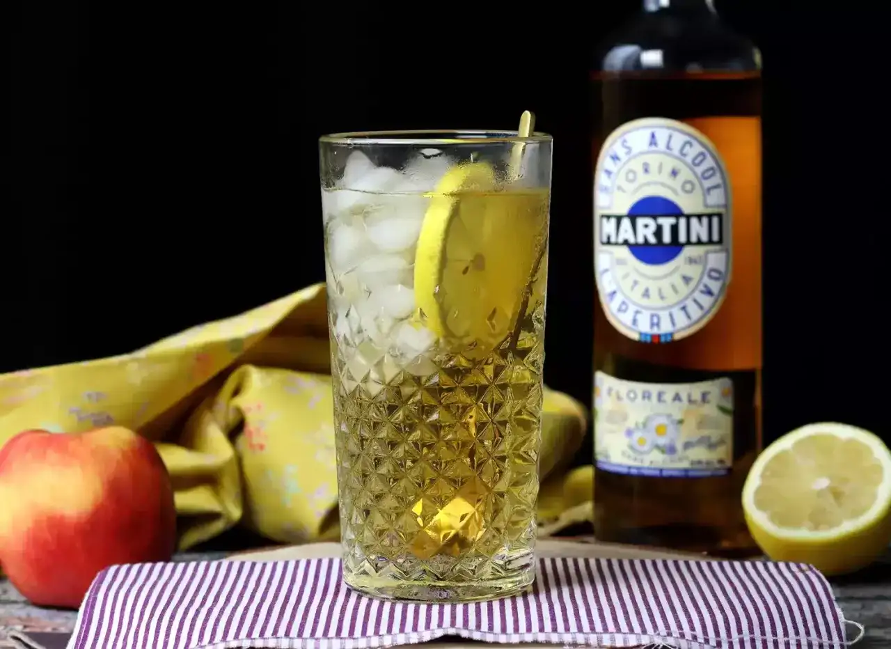 Orzeźwiający spritz bezalkoholowy z plasterkiem cytryny w szklance z lodem. Obok butelka Martini Floreale i jabłko.