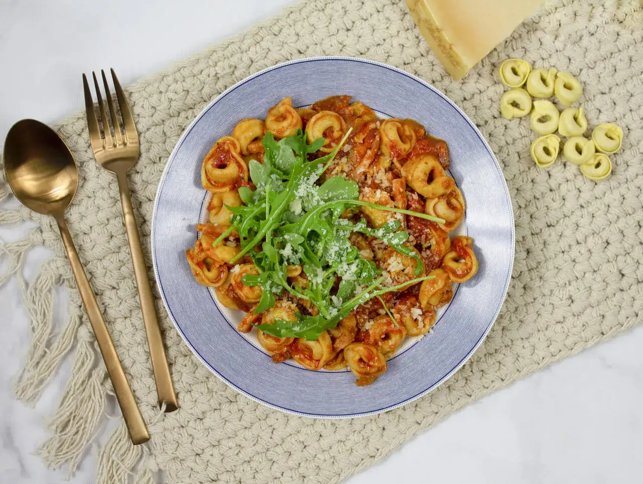 Pyszne tortellini z serem w sosie pomidorowym, udekorowane rukolą i parmezanem. Obok leżą surowe pierożki i kawałek sera.