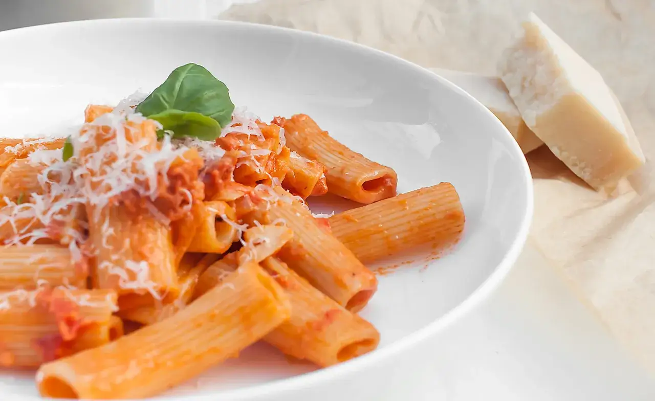 Pyszne mezzi rigatoni w sosie pomidorowym, posypane parmezanem i udekorowane listkiem bazylii. Obok leżą kawałki sera.