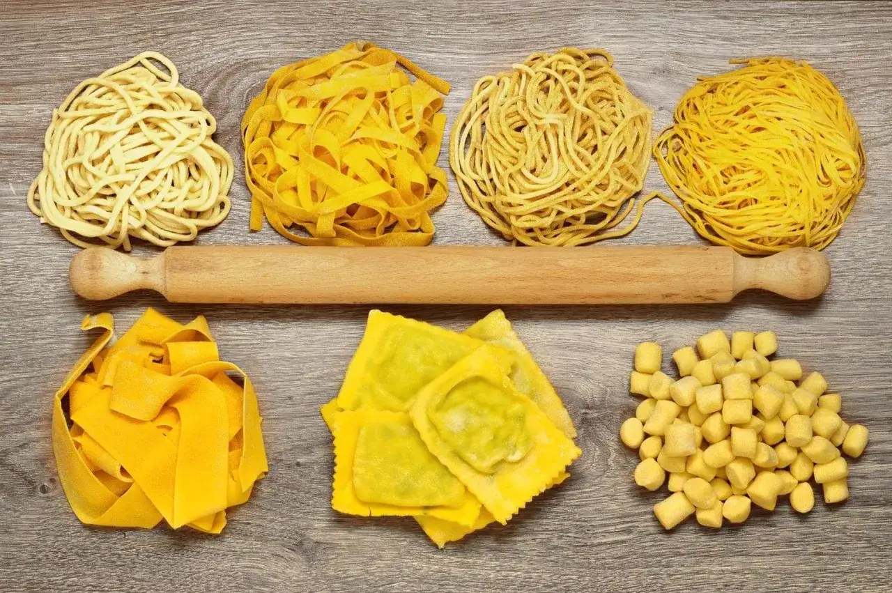 Różne rodzaje makaronów: cienkie nitki, gruby makaron spaghetti, tagliatelle, ravioli i małe kluseczki. Obok wałek do ciasta.