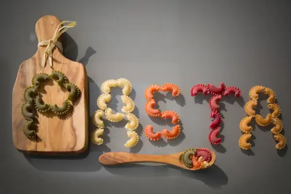 Kolorowy makaron ułożony w napis "PASTA". Kto wymyślił makaron? Może ten, kto ułożył te pyszne kształty na desce i łyżce.