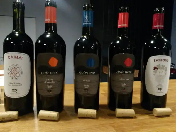 Pięć butelek wina, w tym dwie z etykietą "note nere nero d'avola sicilia doc", stoją na drewnianym stole.