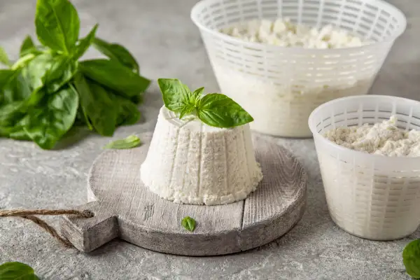 Jak wygląda ser ricotta: świeży, biały serek w kształcie stożka, ozdobiony listkiem bazylii, obok naczynia z serem i gałązka bazylii.