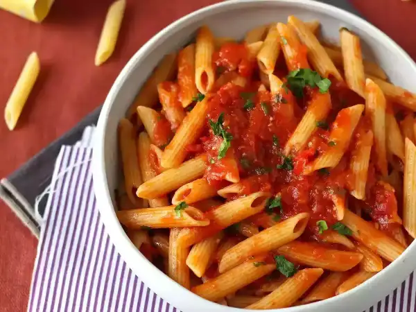 Penne all'arrabbiata w białej misce, polane pikantnym sosem pomidorowym z natką pietruszki. Obok leżą suche makarony.