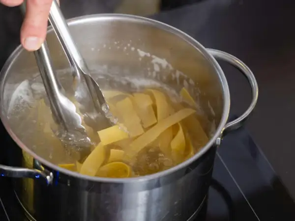 Szczypce wyjmują z wrzącej wody szerokie wstążki makaronu z pszenicy durum, gotowe do podania.