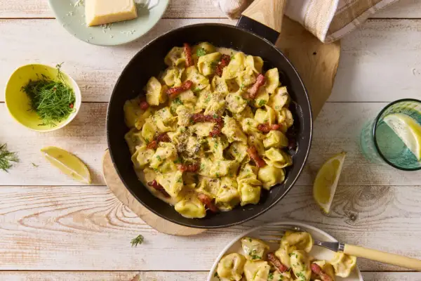 Pyszne tortellini grzybowe w kremowym sosie, posypane parmezanem i boczkiem, podane z cytryną i koperkiem.