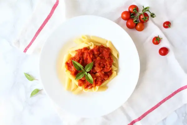 Penne pomodoro z bazylią na białym talerzu, obok gałązka pomidorków koktajlowych.