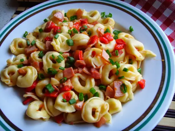 Pyszne tortellini co to jest? Makaron z kawałkami szynki, papryki i szczypiorku na talerzu.