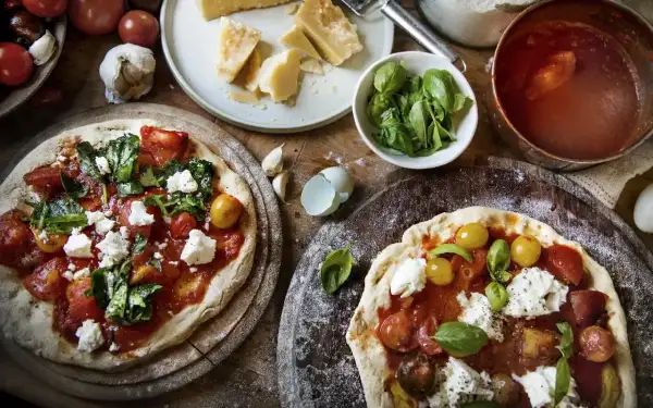 Dwie pizze z sosem pomidorowym, świeżymi pomidorkami, bazylią i kremową włoską mozzarellą. Obok składniki: parmezan, czosnek, pomidory.