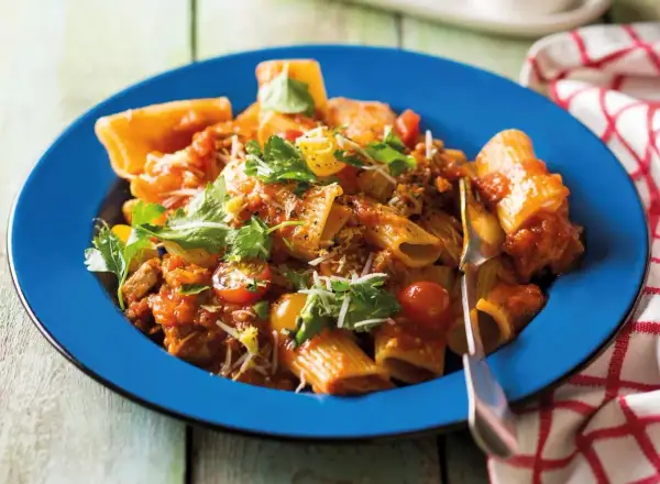 Pyszne mezzi rigatoni w sosie pomidorowym z mięsem mielonym, pomidorkami koktajlowymi i świeżą natką pietruszki, podane na niebieskim talerzu.