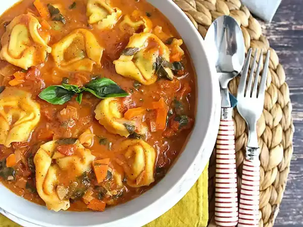 Pyszne tortellini z mięsem w kremowym sosie pomidorowym z warzywami, udekorowane listkiem bazylii. Obok widelec i łyżka.