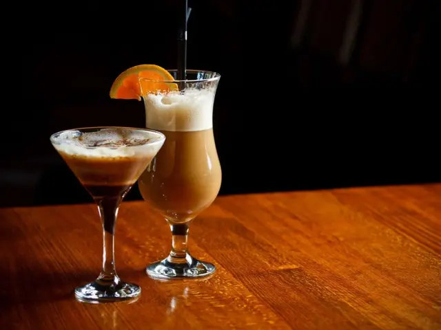 Dwa drinki na drewnianym stole: w kieliszku martini espresso martini, a w wysokiej szklance kawa z alkoholem jak się nazywa, ozdobiona plasterkiem pomarańczy.